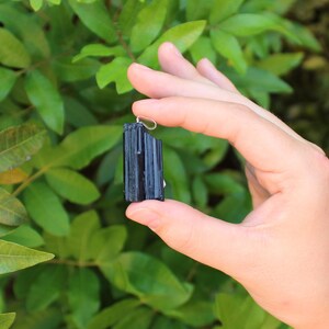 May include: A black tourmaline pendant with a silver-colored bail. The raw, natural crystal is held against a backdrop of green foliage. The tourmaline has a textured surface and a rectangular shape.