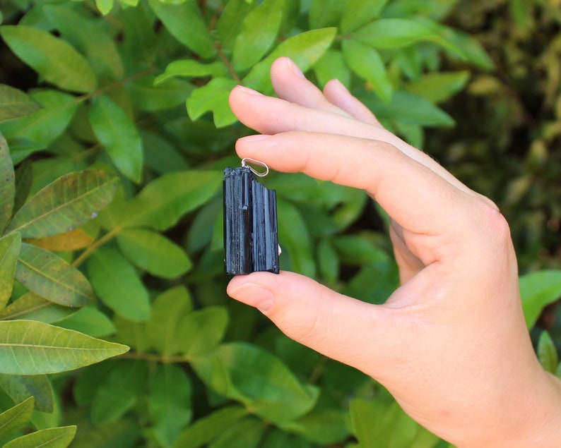 May include: A black tourmaline pendant with a silver-colored bail. The raw, natural crystal is held against a backdrop of green foliage. The tourmaline has a textured surface and a rectangular shape.