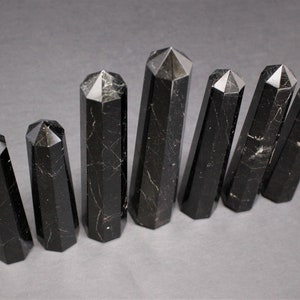 May include: Seven black obsidian crystal points of varying sizes, arranged in a row on a gray surface. The points are all faceted and have a smooth, polished finish.