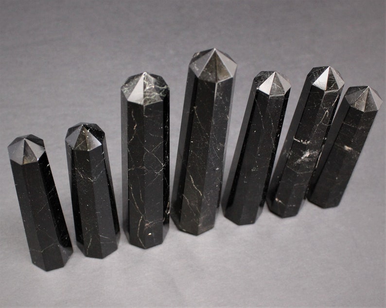 May include: Seven black obsidian crystal points of varying sizes, arranged in a row on a gray surface. The points are all faceted and have a smooth, polished finish.