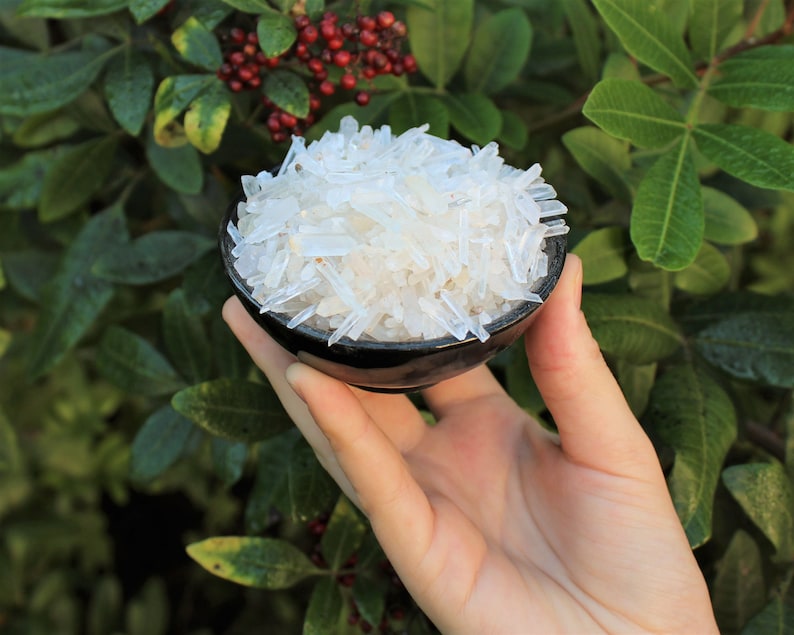 May include: A small black bowl filled with clear, white, crystal-like pieces. The bowl is being held in someone's hand.