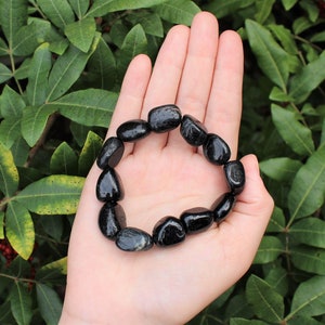 May include: A stretch bracelet composed of smooth, polished black tourmaline gemstone beads. The beads are irregular, rounded, and glossy, strung together in a circular loop to form a continuous band that fits around the wrist.