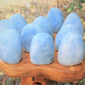 May include: A collection of ten smooth, polished, light blue calcite stones with a crystalline texture. Each stone has an oval, rounded shape and a translucent appearance, displaying variations in color intensity and internal mineral patterns.