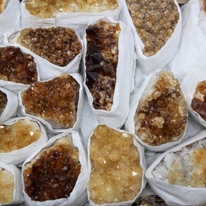 May include: A collection of raw, unpolished citrine crystals wrapped in white tissue paper. The crystals are various shades of yellow and brown, with some having a more translucent appearance.