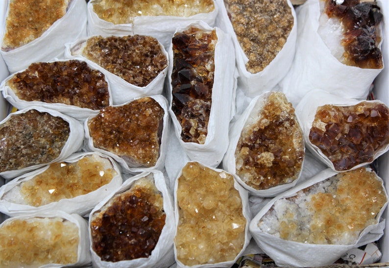 May include: A collection of raw, unpolished citrine crystals wrapped in white tissue paper. The crystals are various shades of yellow and brown, with some having a more translucent appearance.