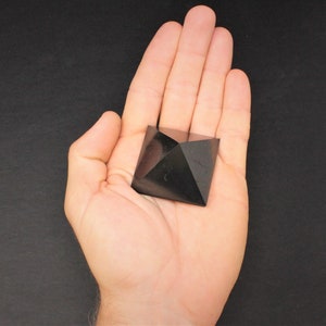 May include: Black obsidian pyramid crystal, a dark, geometric shape, resting in a hand.