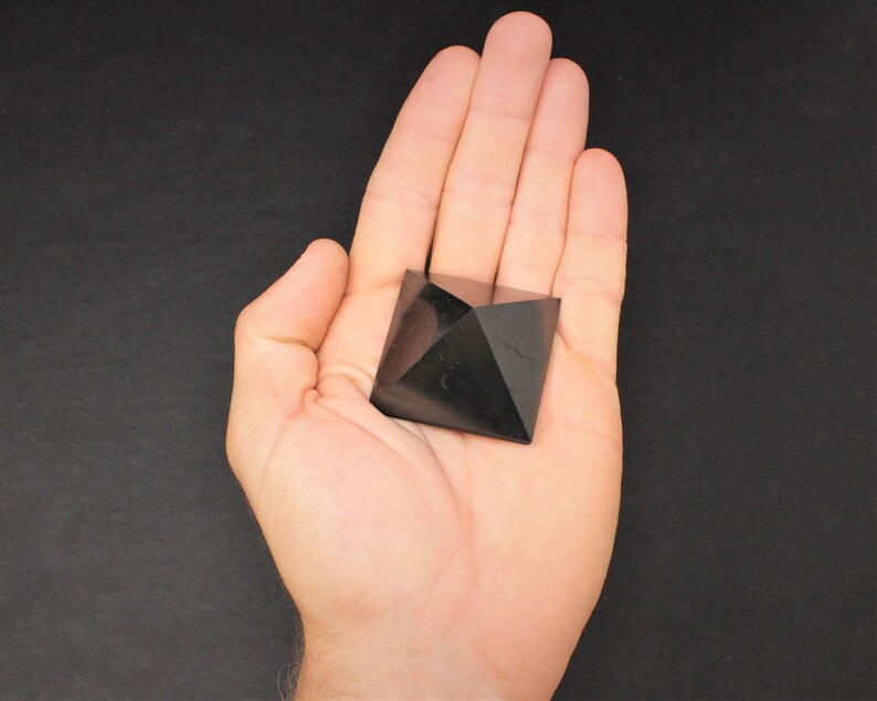 May include: Black obsidian pyramid crystal, a dark, geometric shape, resting in a hand.