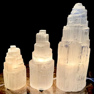 May include: Three selenite crystal lamps of varying sizes, each with a unique, layered tower design. The lamps emit a soft, warm glow, creating a calming ambiance. The largest lamp is approximately 12 inches tall.