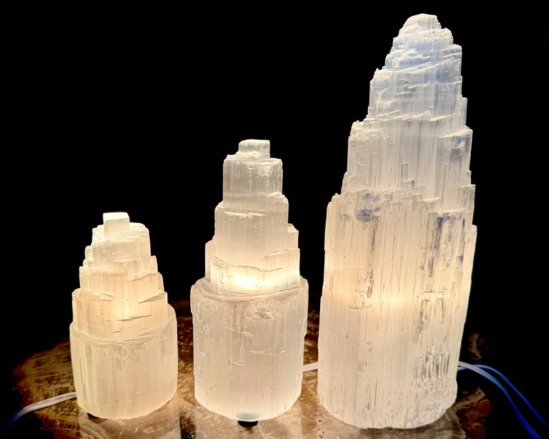May include: Three selenite crystal lamps of varying sizes, each with a unique, layered tower design. The lamps emit a soft, warm glow, creating a calming ambiance. The largest lamp is approximately 12 inches tall.