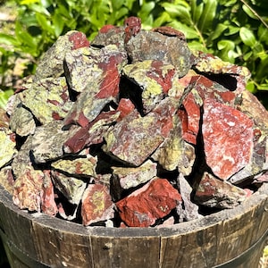 May include: A pile of red and green jasper stones in a wooden bucket. The stones are rough and have a natural, earthy look.
