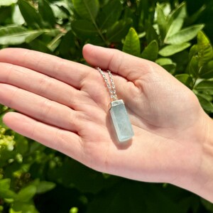 May include: A light blue aquamarine pendant necklace with a silver chain. The rectangular pendant is a pale blue color and is suspended from a delicate silver chain. The necklace is displayed against a background of green foliage.