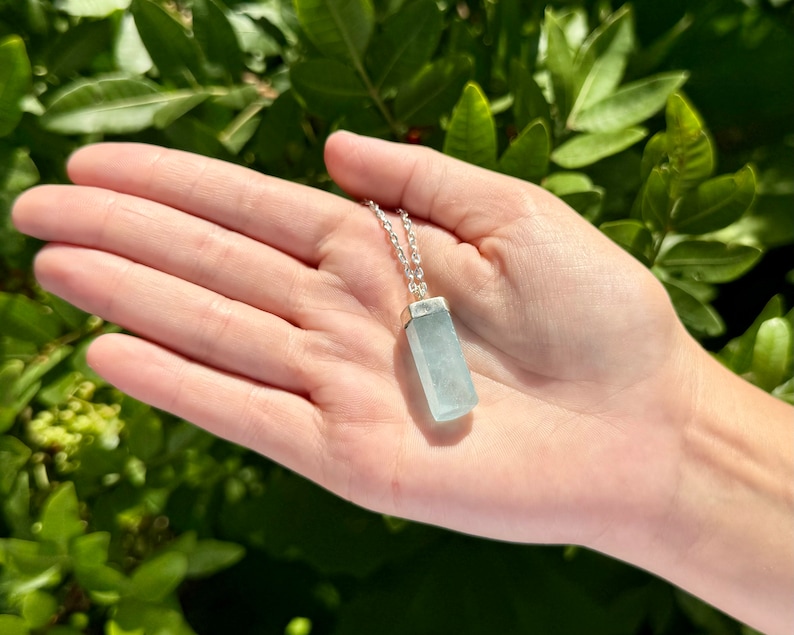 May include: A light blue aquamarine pendant necklace with a silver chain. The rectangular pendant is a pale blue color and is suspended from a delicate silver chain. The necklace is displayed against a background of green foliage.