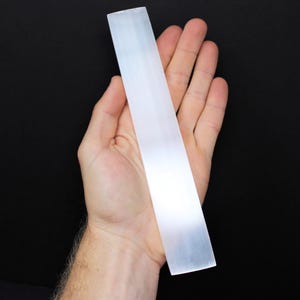 May include: A white selenite wand, also known as a cleansing wand, is held in a hand against a black background.