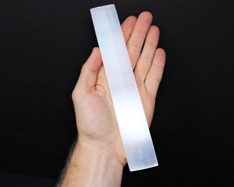 May include: A white selenite wand, also known as a cleansing wand, is held in a hand against a black background.