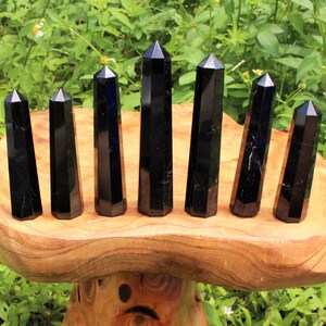 May include: Seven black obsidian crystal points of varying sizes are arranged in a row on a wooden stand. The points are all faceted and have a smooth, polished finish.