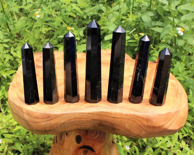 May include: Seven black obsidian crystal points of varying sizes are arranged in a row on a wooden stand. The points are all faceted and have a smooth, polished finish.