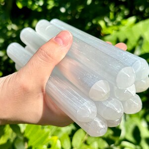 May include: A bundle of selenite wands, also known as satin spar, in a hand. The wands are white and have a smooth, polished surface. They are approximately 4-5 inches long.