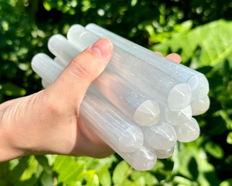 May include: A bundle of selenite wands, also known as satin spar, in a hand. The wands are white and have a smooth, polished surface. They are approximately 4-5 inches long.
