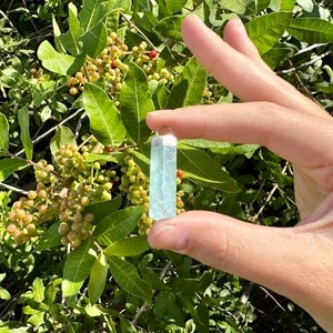 May include: A rectangular, light blue aquamarine pendant with a silver-colored cap. The gemstone is held against a backdrop of green leaves and berries. The pendant is approximately 1.5 inches in length.