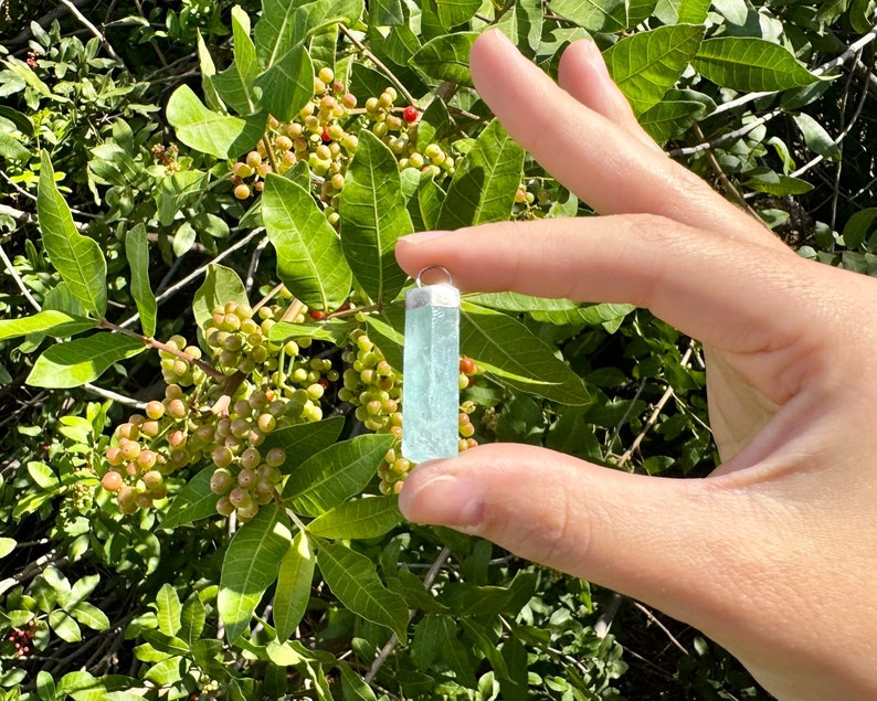 May include: A rectangular, light blue aquamarine pendant with a silver-colored cap. The gemstone is held against a backdrop of green leaves and berries. The pendant is approximately 1.5 inches in length.