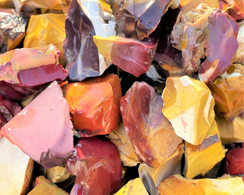 May include: A collection of raw, uncut gemstones in various colors. The stones display a range of hues, including deep reds, oranges, yellows, and pinks. The surfaces of the stones are rough and textured, with a natural, unpolished appearance.