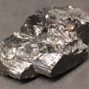 May include: A close-up of a dark, metallic-looking mineral specimen. The surface is highly reflective with a fractured, crystalline structure. The color is primarily a deep gray with hints of silver and black, and some reddish-brown inclusions.