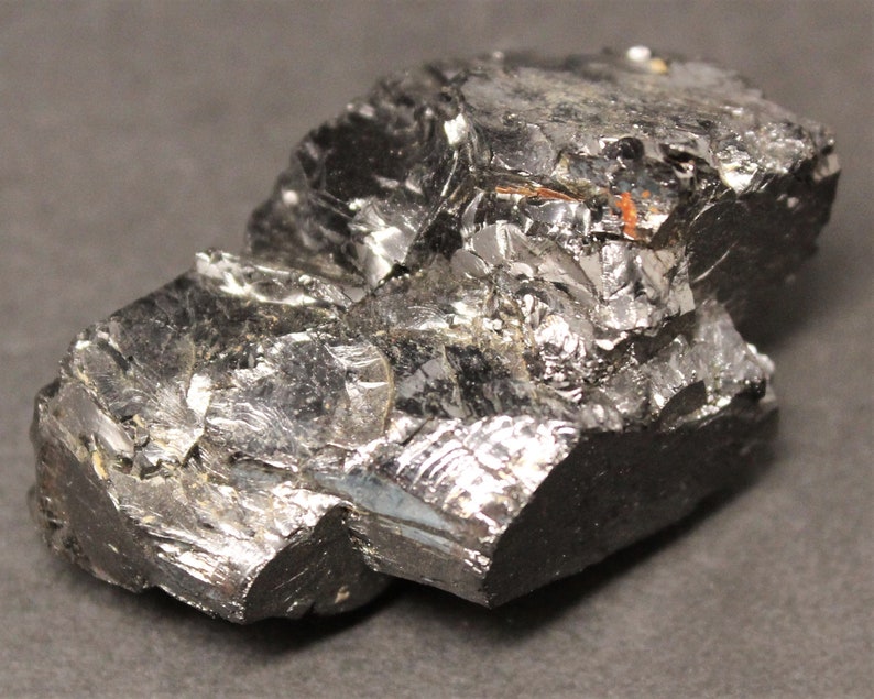 May include: A close-up of a dark, metallic-looking mineral specimen. The surface is highly reflective with a fractured, crystalline structure. The color is primarily a deep gray with hints of silver and black, and some reddish-brown inclusions.