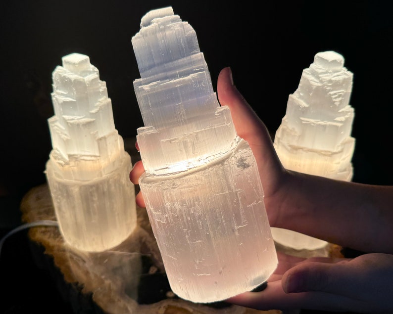 May include: Three white selenite lamps with a layered, stacked design. The lamps are illuminated from within, creating a soft glow. The lamps are on a wooden base, with a dark background.