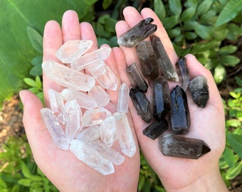 May include: Two hands holding a selection of clear quartz and smoky quartz crystals. The crystals are all pointed and have a natural, rough texture.