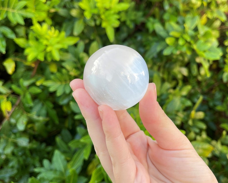 Op de afbeelding: Een witte selenietbol, ook wel een selenietkogel genoemd, wordt in een hand gehouden tegen een groene, bladerrijke achtergrond. De bol is glad en heeft een doorschijnende uitstraling.