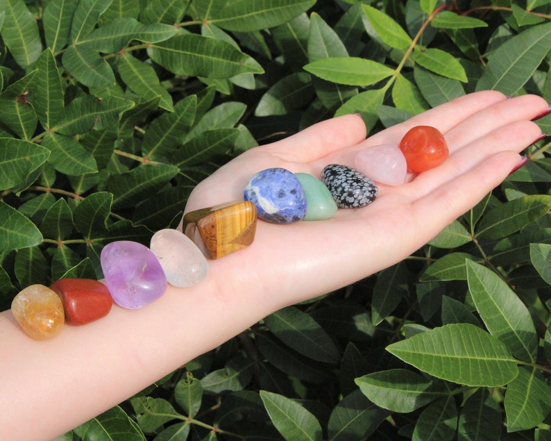 May include: A hand holding seven smooth, polished stones in a rainbow of colors, including red, orange, yellow, green, blue, indigo, and violet. These stones are often used in crystal healing and chakra balancing.