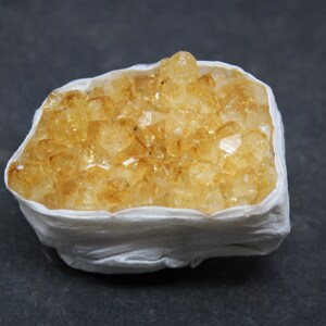 May include: A cluster of yellow citrine crystals in a white paper wrapping. The crystals are clustered together in a natural formation.