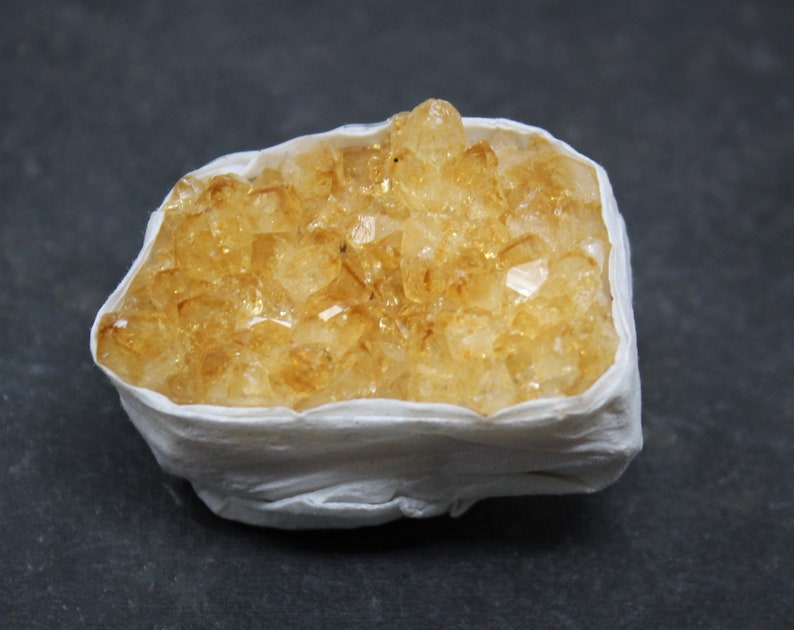 May include: A cluster of yellow citrine crystals in a white paper wrapping. The crystals are clustered together in a natural formation.