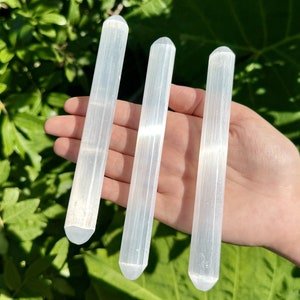 May include: Three white selenite wands, also known as selenite sticks, are held in a hand. The wands are smooth and have a striated texture.