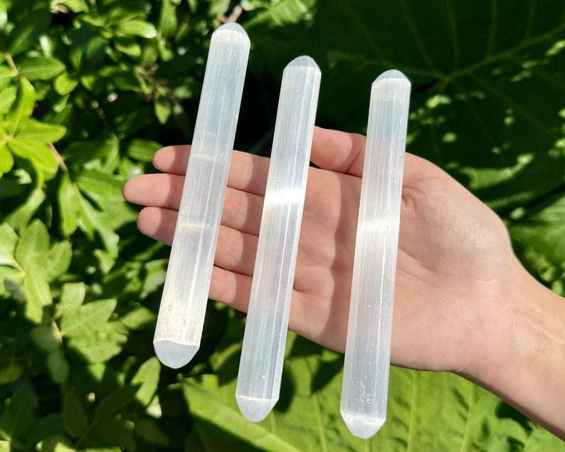 May include: Three white selenite wands, also known as selenite sticks, are held in a hand. The wands are smooth and have a striated texture.