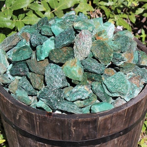 May include: A wooden bucket filled with a large quantity of green aventurine rough stones. The stones are various sizes and shapes, with a deep green color and a sparkly, glittery texture.