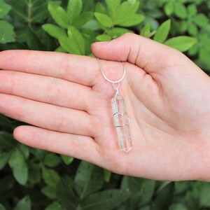 May include: A clear quartz crystal pendant wrapped in silver wire, hanging from a silver chain. The pendant is about 2 inches long and is held in a hand.