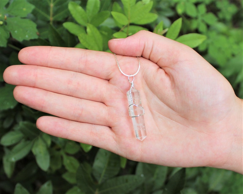 May include: A clear quartz crystal pendant wrapped in silver wire, hanging from a silver chain. The pendant is about 2 inches long and is held in a hand.
