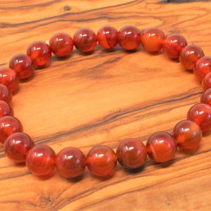 May include: A beaded bracelet featuring polished, round beads in shades of red and orange. The beads are strung together to form a circular shape. The bracelet is displayed on a wooden surface.
