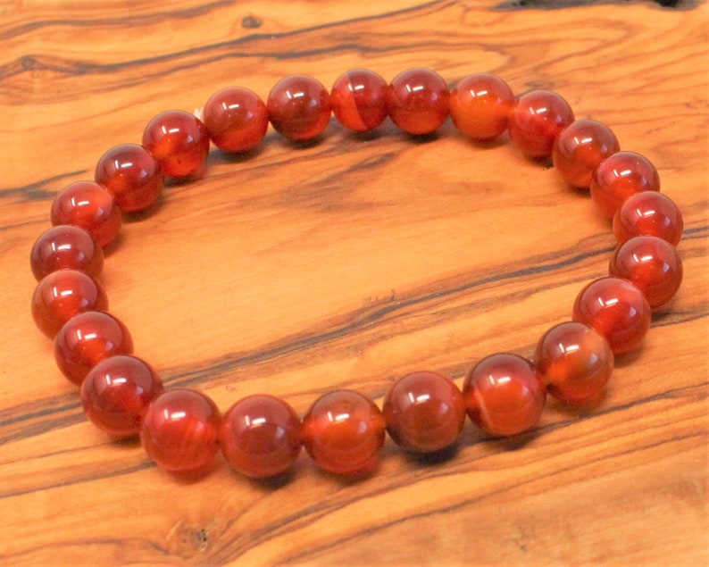 May include: A beaded bracelet featuring polished, round beads in shades of red and orange. The beads are strung together to form a circular shape. The bracelet is displayed on a wooden surface.