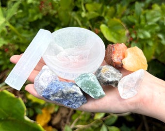 May include: A white selenite bowl and a long selenite wand accompanied by a collection of raw crystals including blue sodalite, green aventurine, clear quartz, orange calcite, and a dark brown stone with reddish inclusions held in a palm.