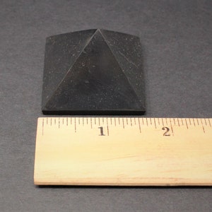 May include: A black obsidian pyramid measuring 1 inch across the base. The pyramid is sitting on a wooden ruler.