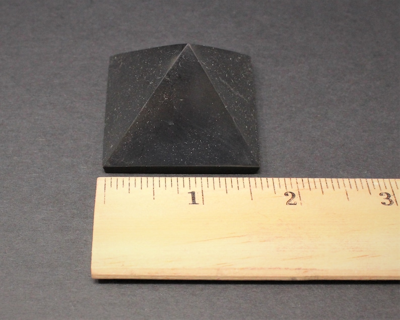 May include: A black obsidian pyramid measuring 1 inch across the base. The pyramid is sitting on a wooden ruler.