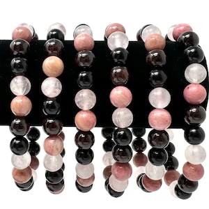 May include: Six bracelets made of black, pink, and white beads. The bracelets are stacked on a black surface.