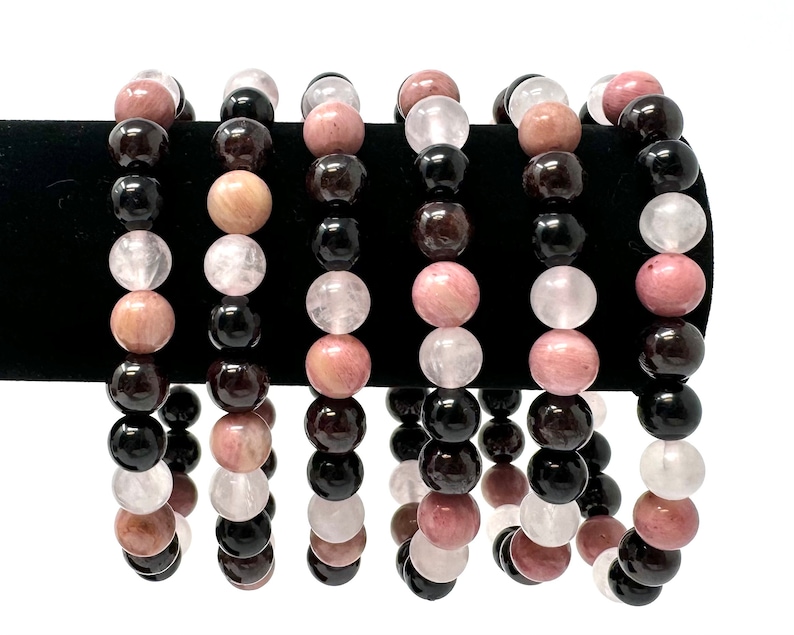 May include: Six bracelets made of black, pink, and white beads. The bracelets are stacked on a black surface.