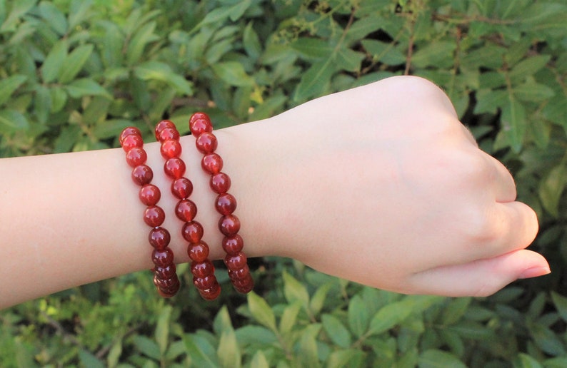 May include: Three red beaded bracelets. Each bracelet is made of small, round, reddish-brown beads. The bracelets are worn together on a wrist, with a green leafy background.