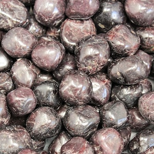 May include: A close-up of a pile of tumbled red-brown garnet stones. The stones are smooth and polished, with a glossy finish.