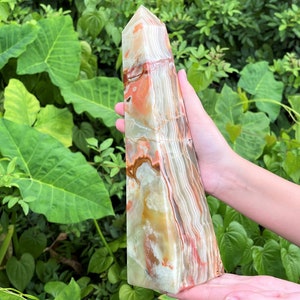 May include: A polished stone tower with a pointed top, featuring a banded pattern of translucent white, cream, and vibrant orange-red mineral layers. The crystal obelisk displays natural veining and smooth, glossy surfaces throughout its structure.