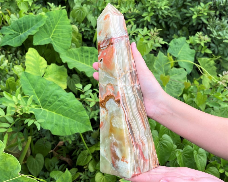 May include: A polished stone tower with a pointed top, featuring a banded pattern of translucent white, cream, and vibrant orange-red mineral layers. The crystal obelisk displays natural veining and smooth, glossy surfaces throughout its structure.