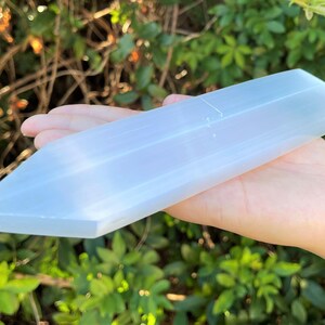 May include: A long, translucent white selenite crystal wand with a smooth, polished surface and a pointed tip. The crystal has a rectangular, faceted shape with a soft, milky appearance that catches the light, showcasing its natural fibrous texture.